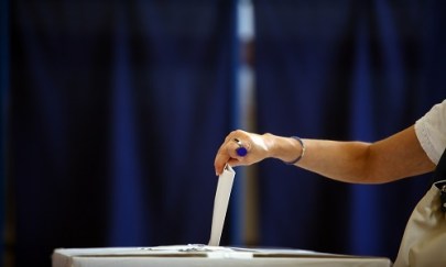 Picture of a hand dropping a ballot into a voting box. A blue curtain is in the backdrop out of focus.