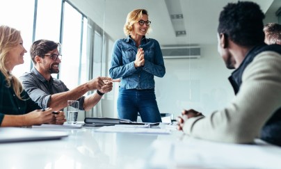 A woman addresses colleagues at the head of a conference table.