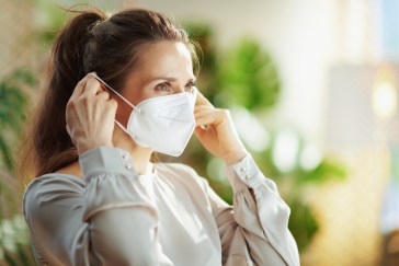 A woman in a grey blouse is putting on a KN95 mask.