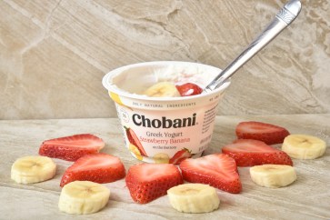 Chobani's strawberry banana yogurt sitting on a table surrounded by strawberry and banana slices.