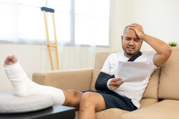man with cast on his foot sits on couch looking at medical bill in stress