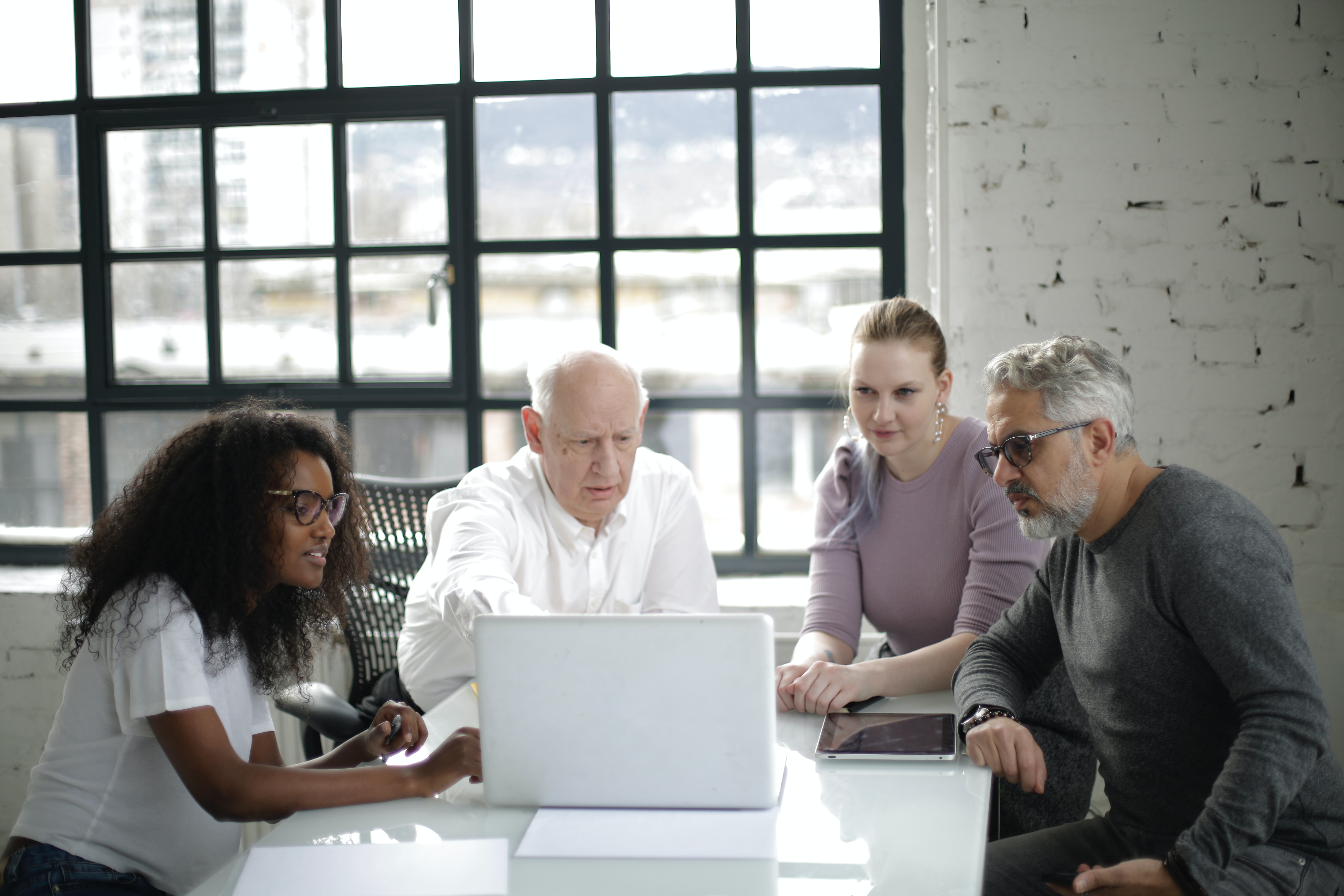 Two young women and two older men sit in an office, all speaking at a laptop, presumably in a group meeting with their virtual colleagues.