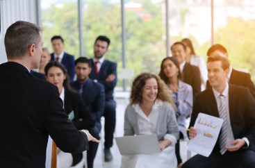 A man is speaking to a crowd of engaged listeners in business clothing.