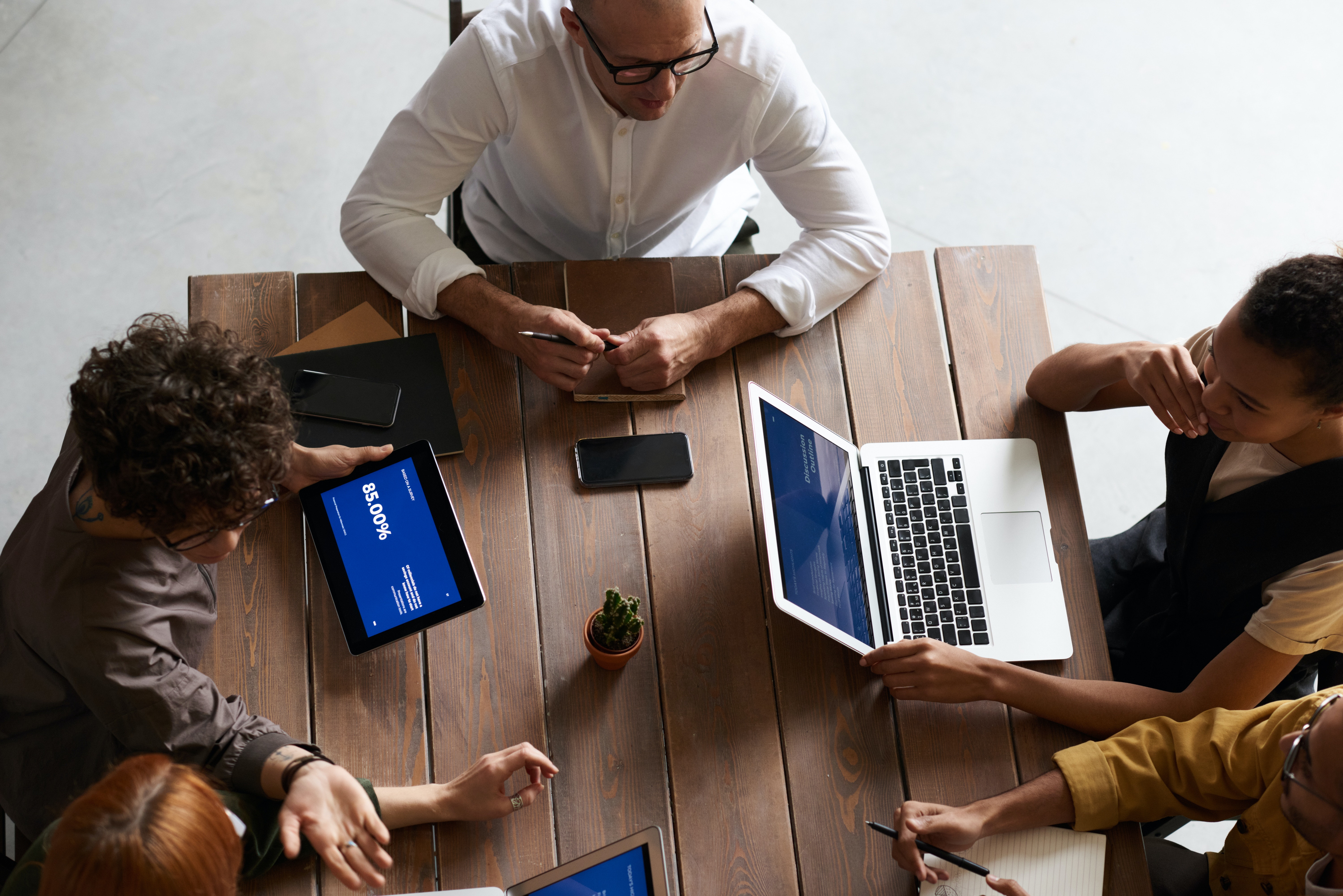 Three team members sit around a conference table, one on an ipad, one on a laptop, and the other taking notes by hand.
