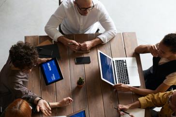 Three team members sit around a conference table, one on an ipad, one on a laptop, and the other taking notes by hand.