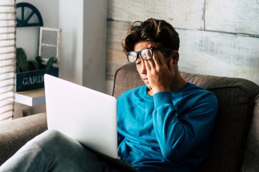 A man rubs his eyes while looking at his laptop.