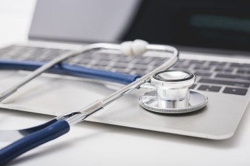 An open Mac laptop with a blue stethoscope resting on the keyboard