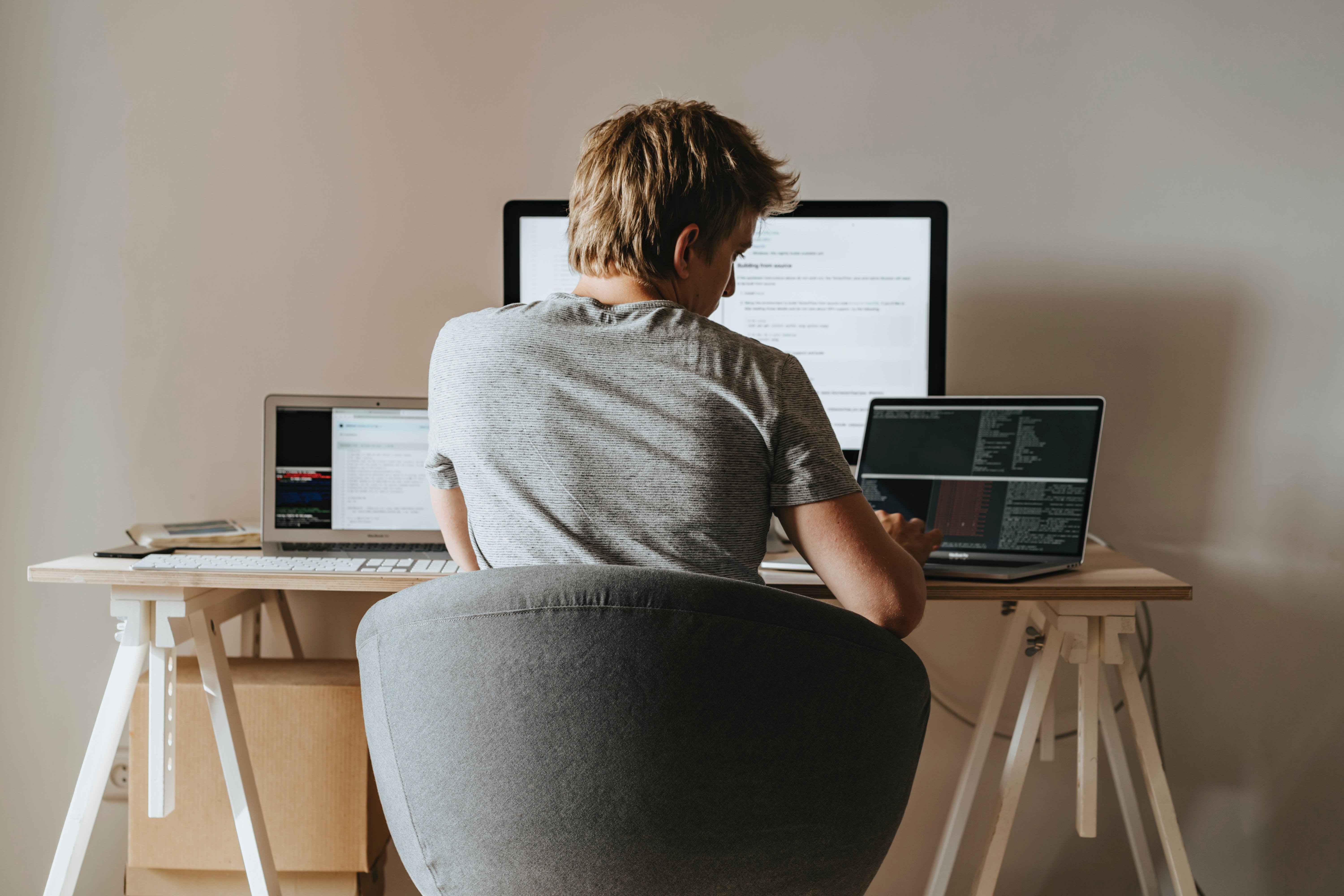A man sits at a desk with three computer monitors. His back is to the reader, and it's implied he's working from home.