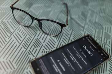A pair of eyeglasses sits on a surface next to a smart phone that has ChatGPT pulled up on its screen.