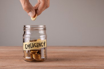 and hand is dropping coins into a mason jar labeled 'emergency' and half full with change