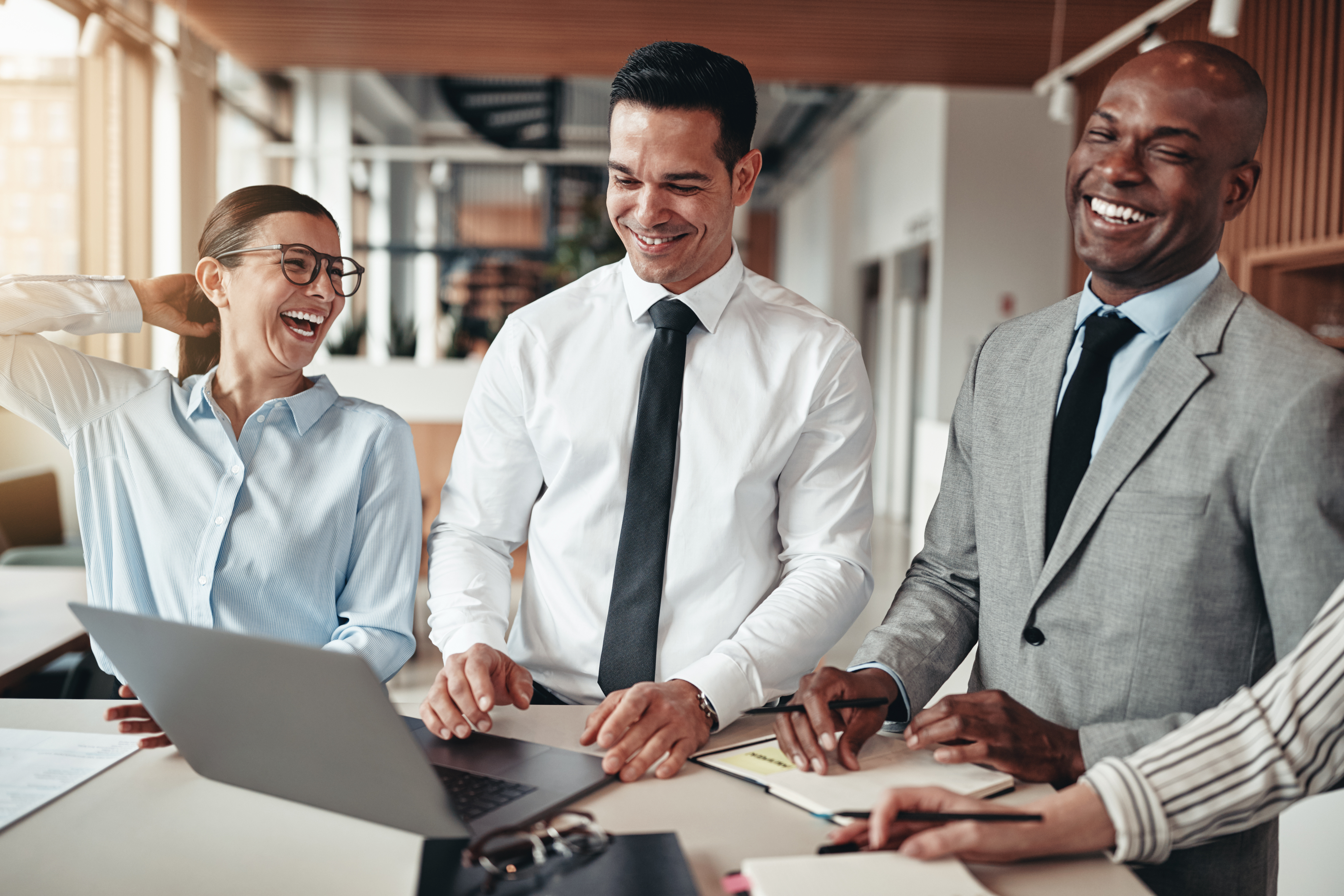 A woman and two men stand around a conference table in business attire, laughing and collaborating.