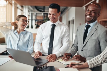 A woman and two men stand around a conference table in business attire, laughing and collaborating.