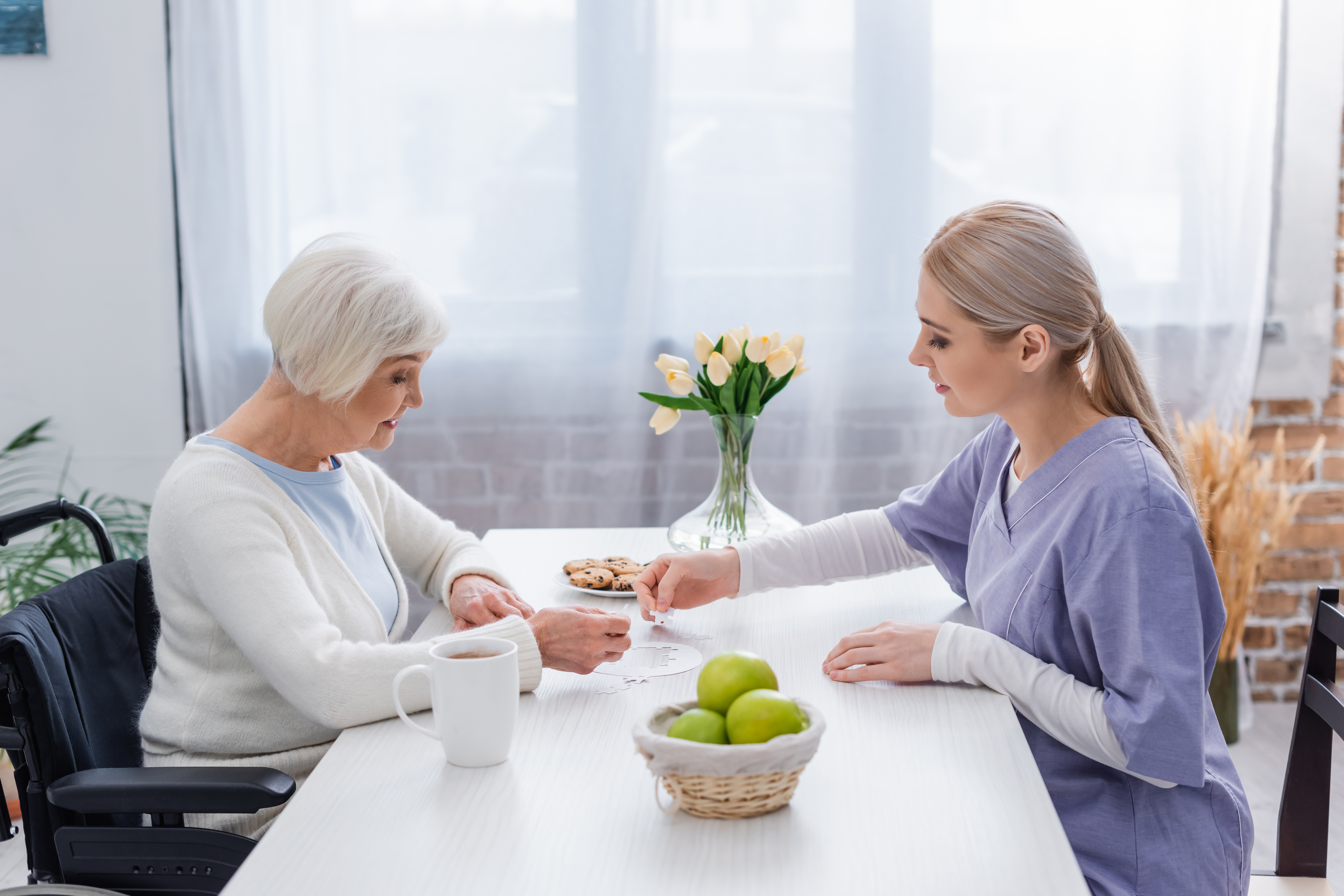 And older woman sits at a table in her home, playing a card game with a female nurse in purple scrubs.