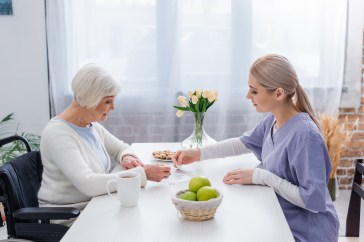 And older woman sits at a table in her home, playing a card game with a female nurse in purple scrubs.