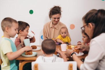 Two daycare teachers sit at a table with an infant and four toddlers