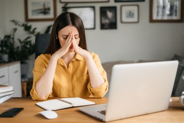 A woman dressed in a yellow blouse, in front of her laptop, looking stressed.