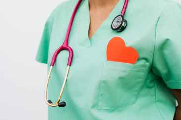 A doctor wearing scrubs and a stethoscope has a red paper heart cutout peeking out of the pocket