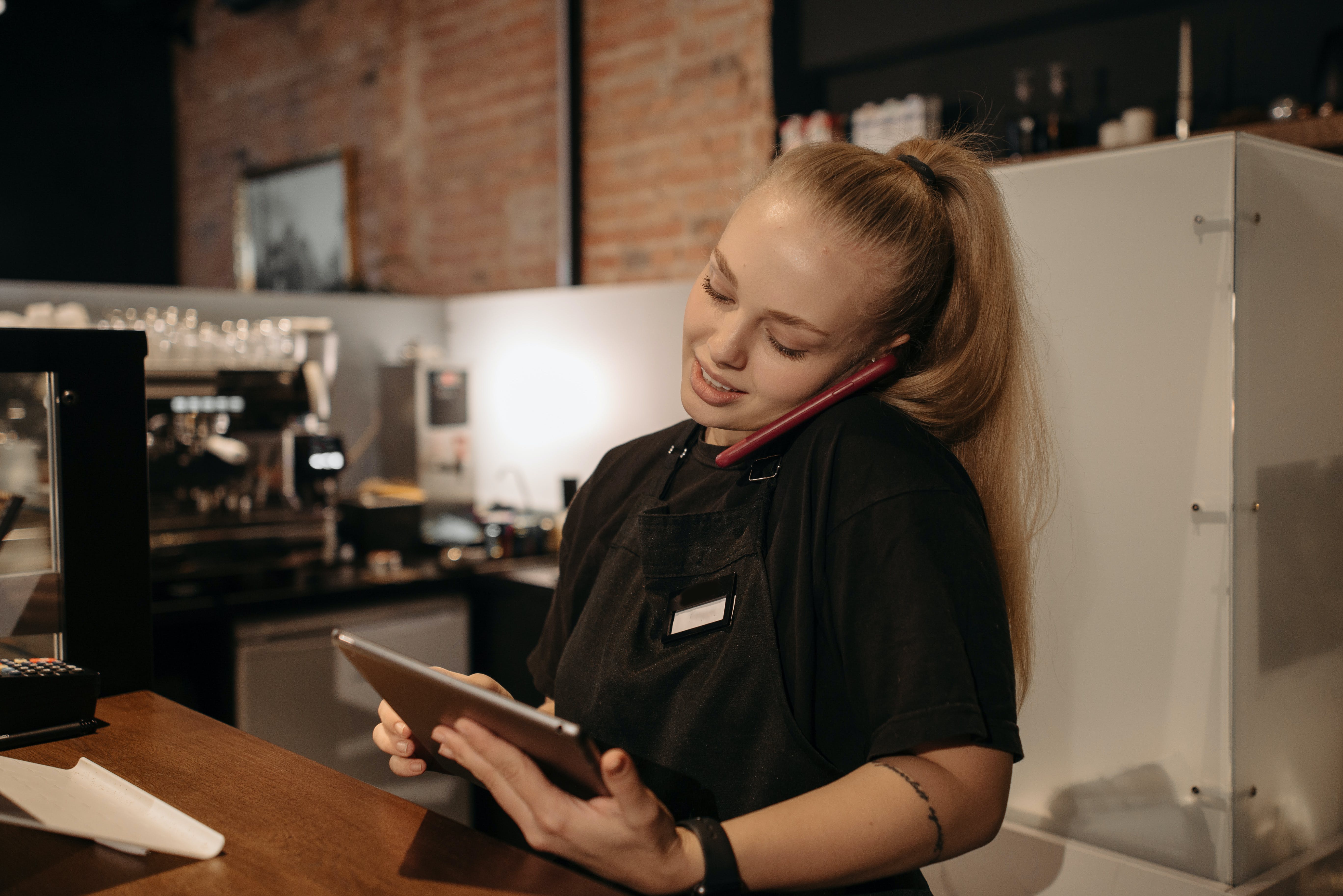 A woman employee stands behind a coffee counter taking an order over the phone and entering it on a tablet.