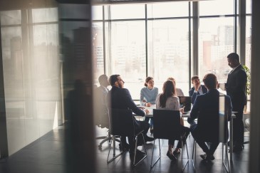 Eight professionals are sitting at a conference table, listening to a colleague speak.