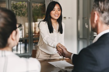 Two managers shake the hand of a young Asian woman in a conference room.