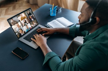 A Black man with a headset is on a video call with colleagues at his desk in his living room.
