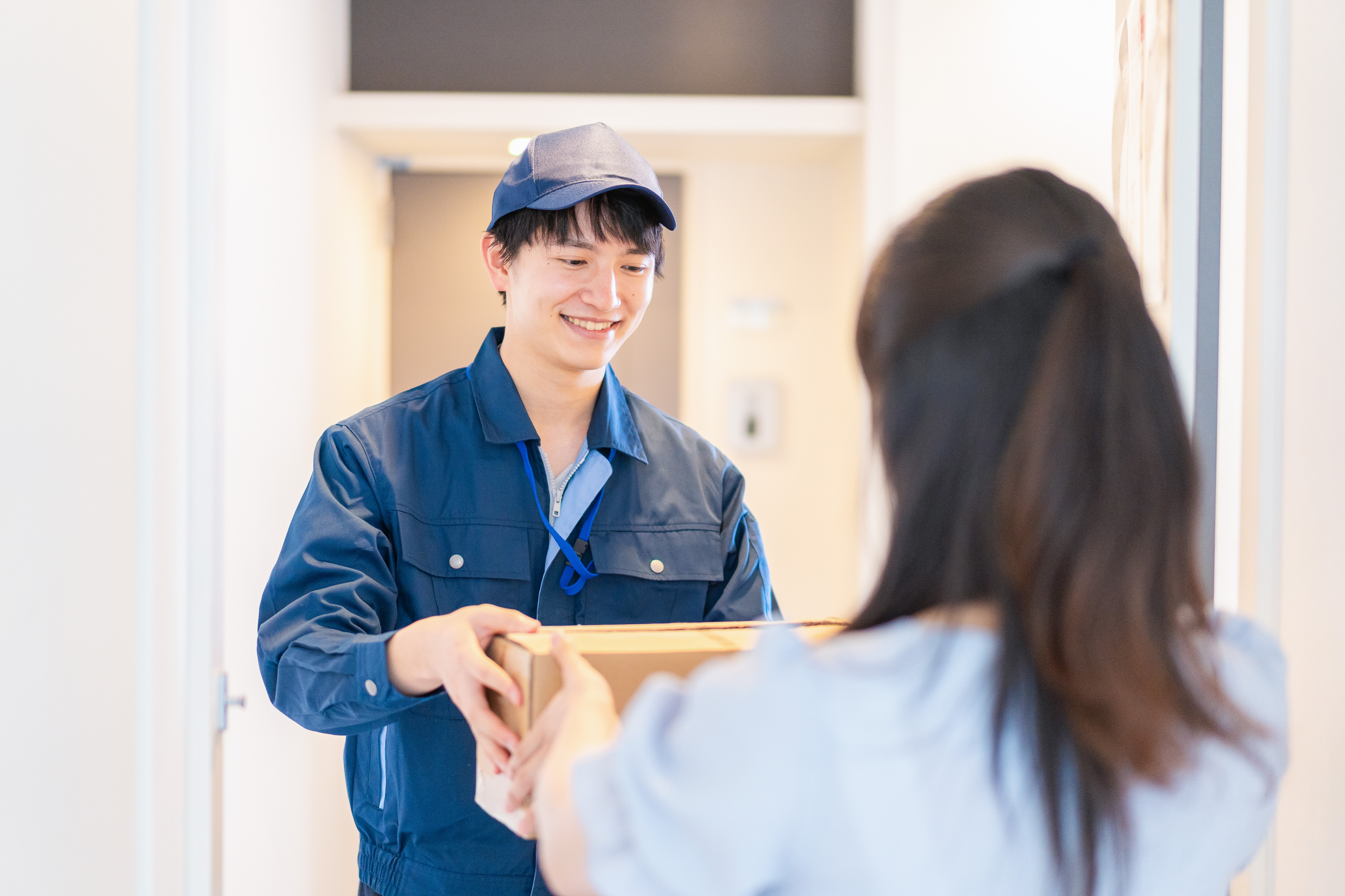 A smiling delivery man hands a brown-box package to a woman at the threshold of her apartment.