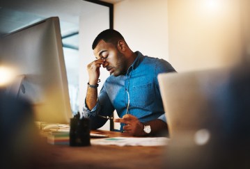 A frustrated Black man pinches the bridge of his nose while facing his desktop computer, glasses in his other hand.