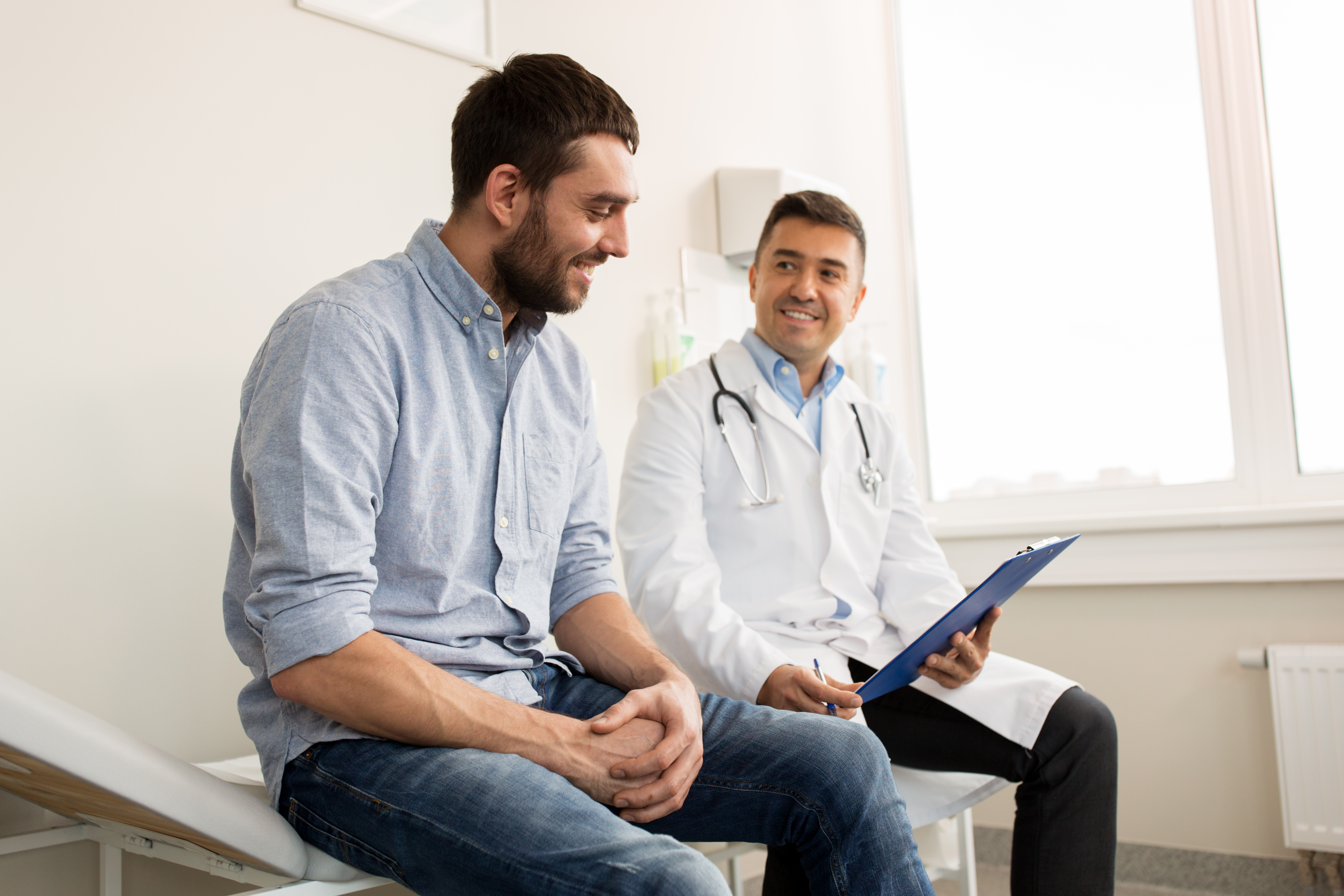 A man in his 40s sits next to a doctor who is holding his patient chart. They are both smiling.