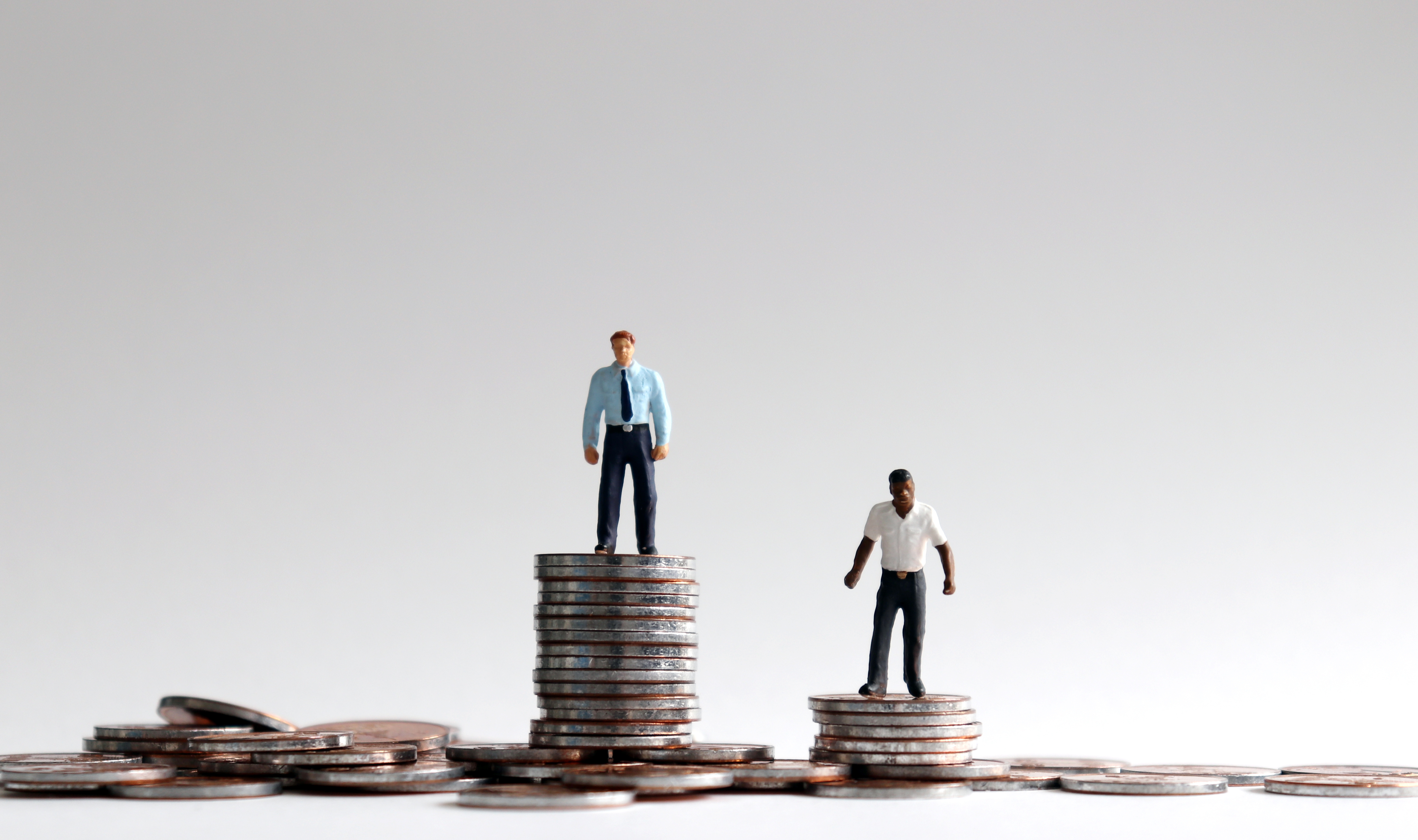 A figurine of a white businessman standing on a stack of quarters, next to a shorter stack of quarters with a black figurine on top