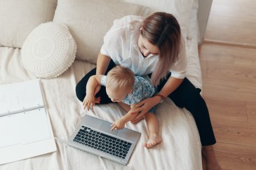Woman sitting on a bed with her baby who is pressing buttons on the computer.
