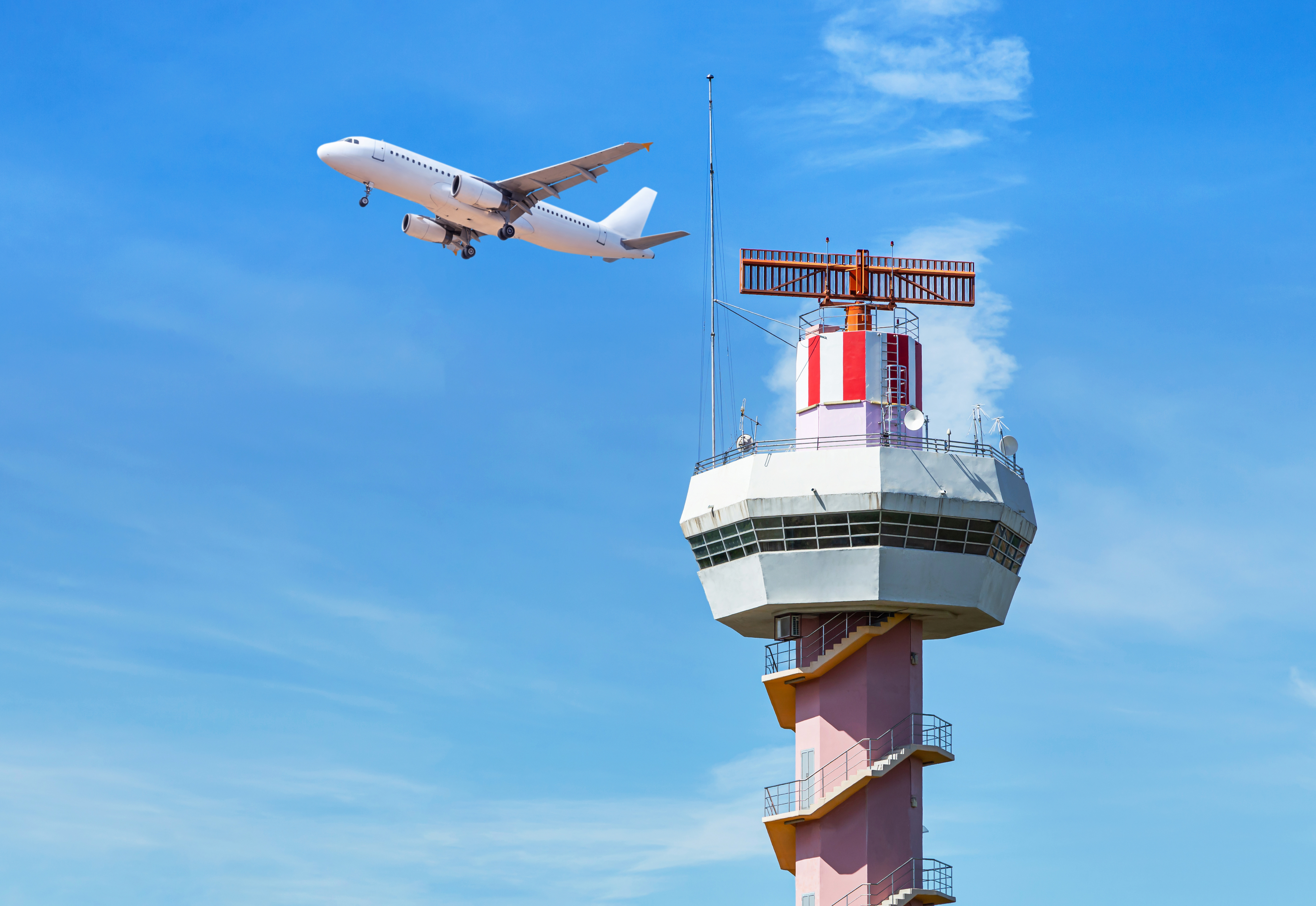 Airport traffic control tower in the foreground, a plane flies behind it, and the sky is blue.