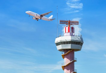 Airport traffic control tower in the foreground, a plane flies behind it, and the sky is blue.