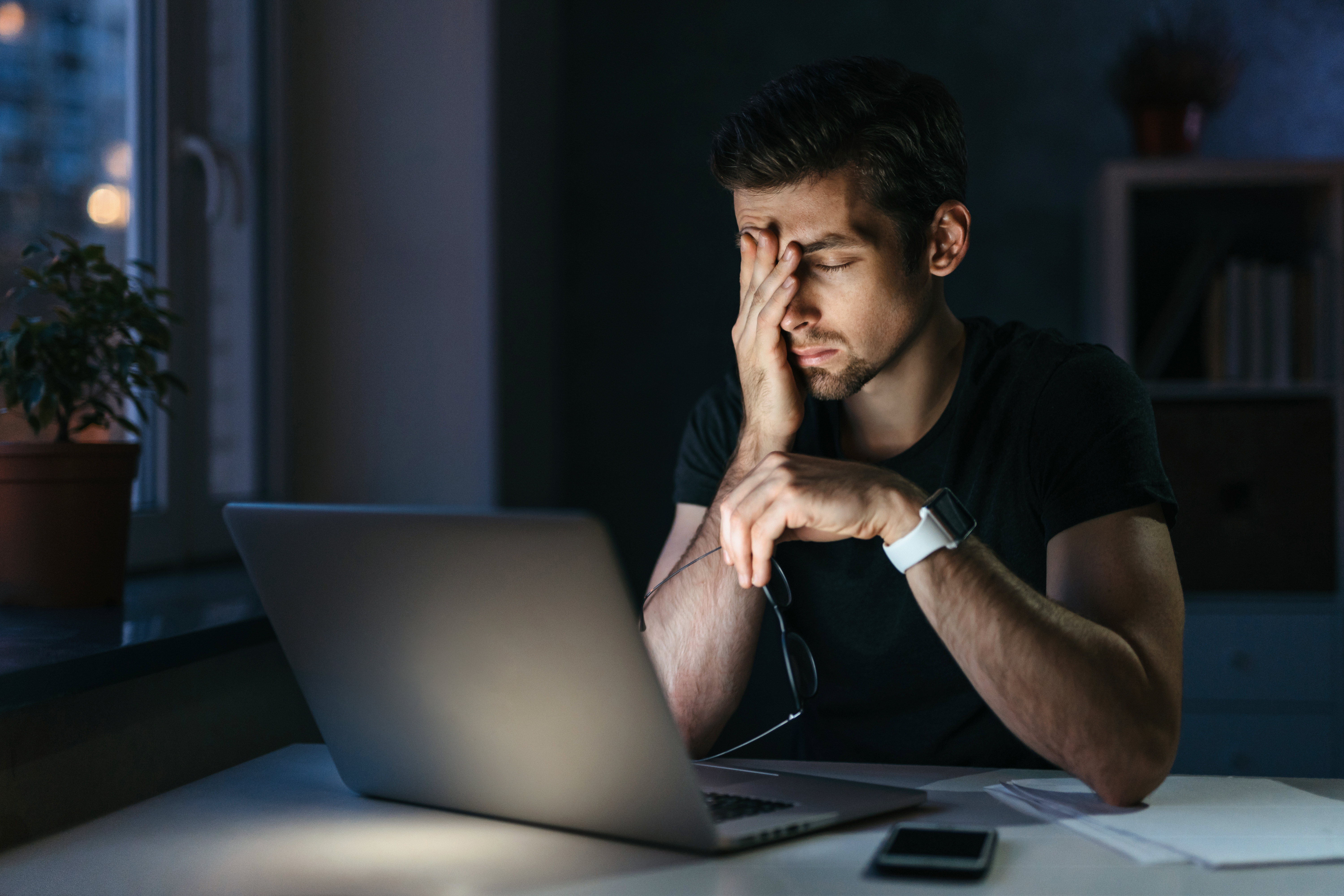 A man rubs his face with his glasses in the other hand in front of his laptop; it's nighttime, and there's a soft glow from the laptop.