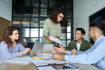 Four colleagues of different ethnicities discuss something on pieces of paper laid around a wooden conference table.