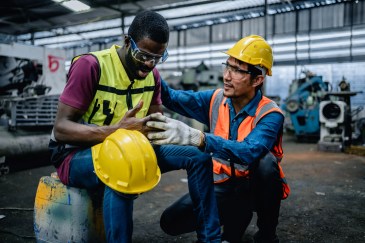 Two construction workers in yellow hard hats; one worker injured his finger, and the other is kneeling next to him.