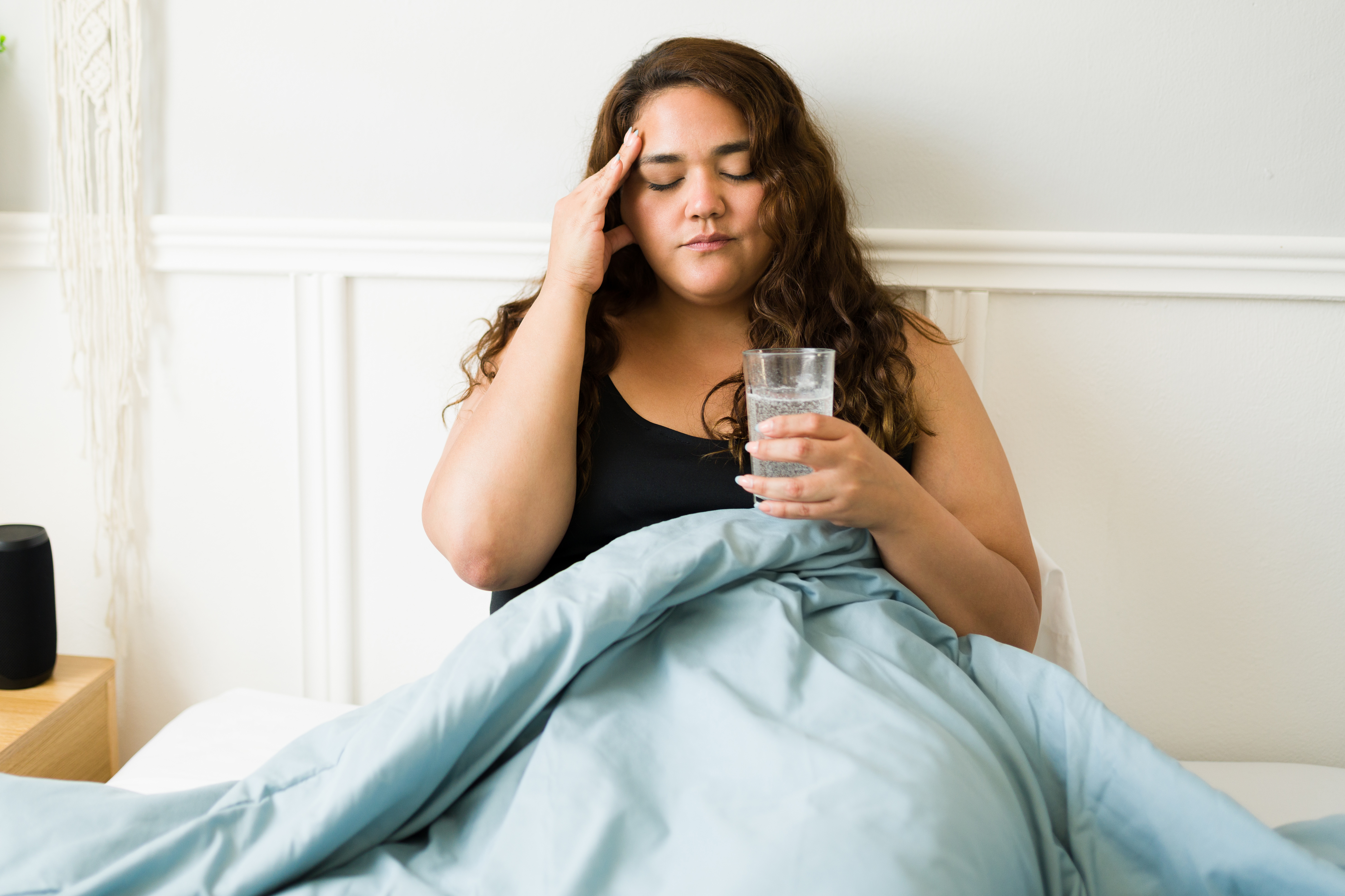 A woman with long, curly brown hair is leaning against the bed headboard, holding a glass of water and rubbing her temple.