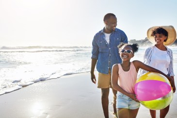 A Black family with a young daughter walks along the beach; the daughter has a yellow and pink beach ball, and she's laughing.