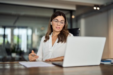 A woman with brown hair and glasses, speaking at her white laptop with a pen in her hand.
