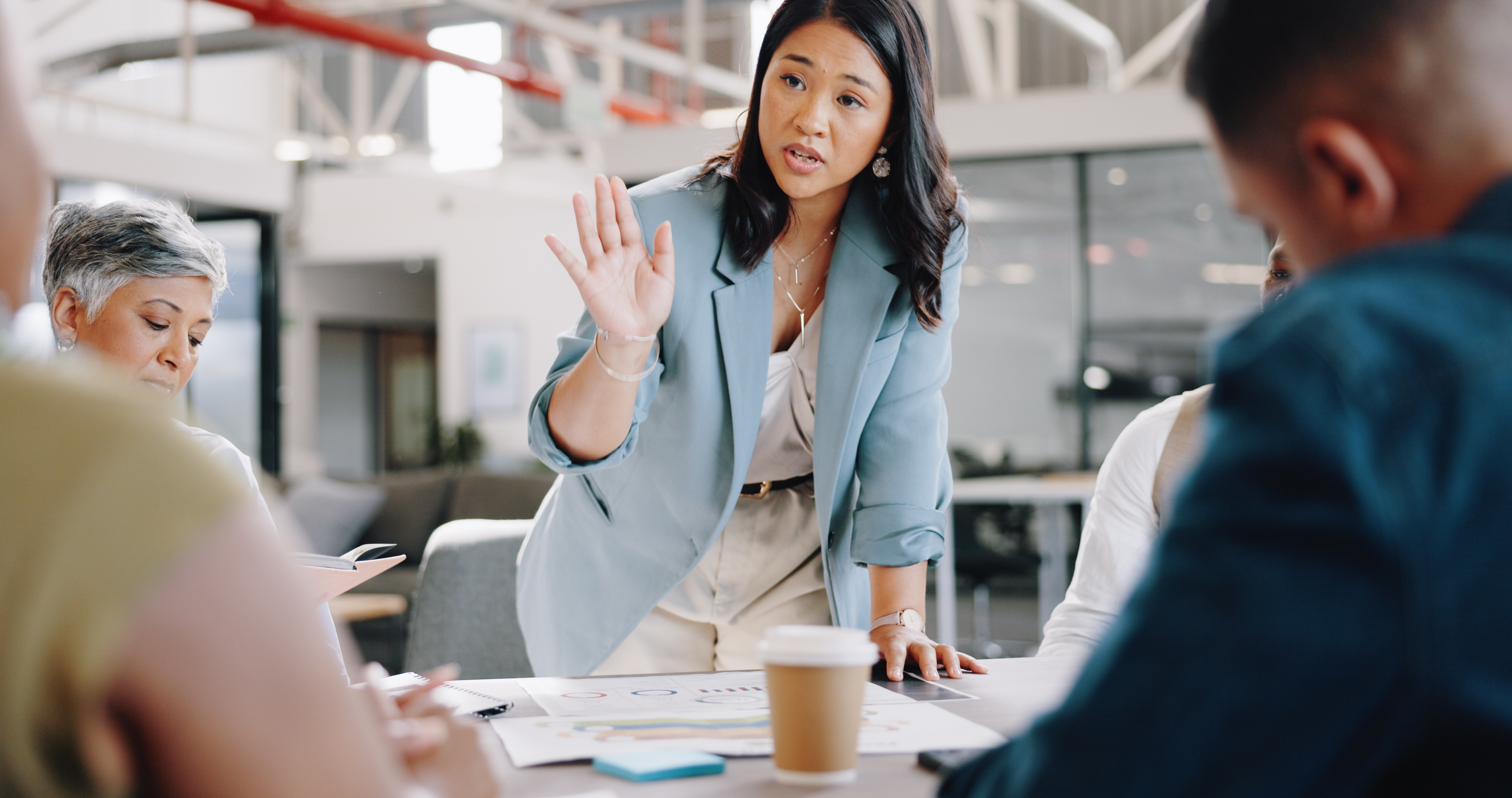 An Asian woman with a light blue blazer stands over a conference table, addressing four colleagues.