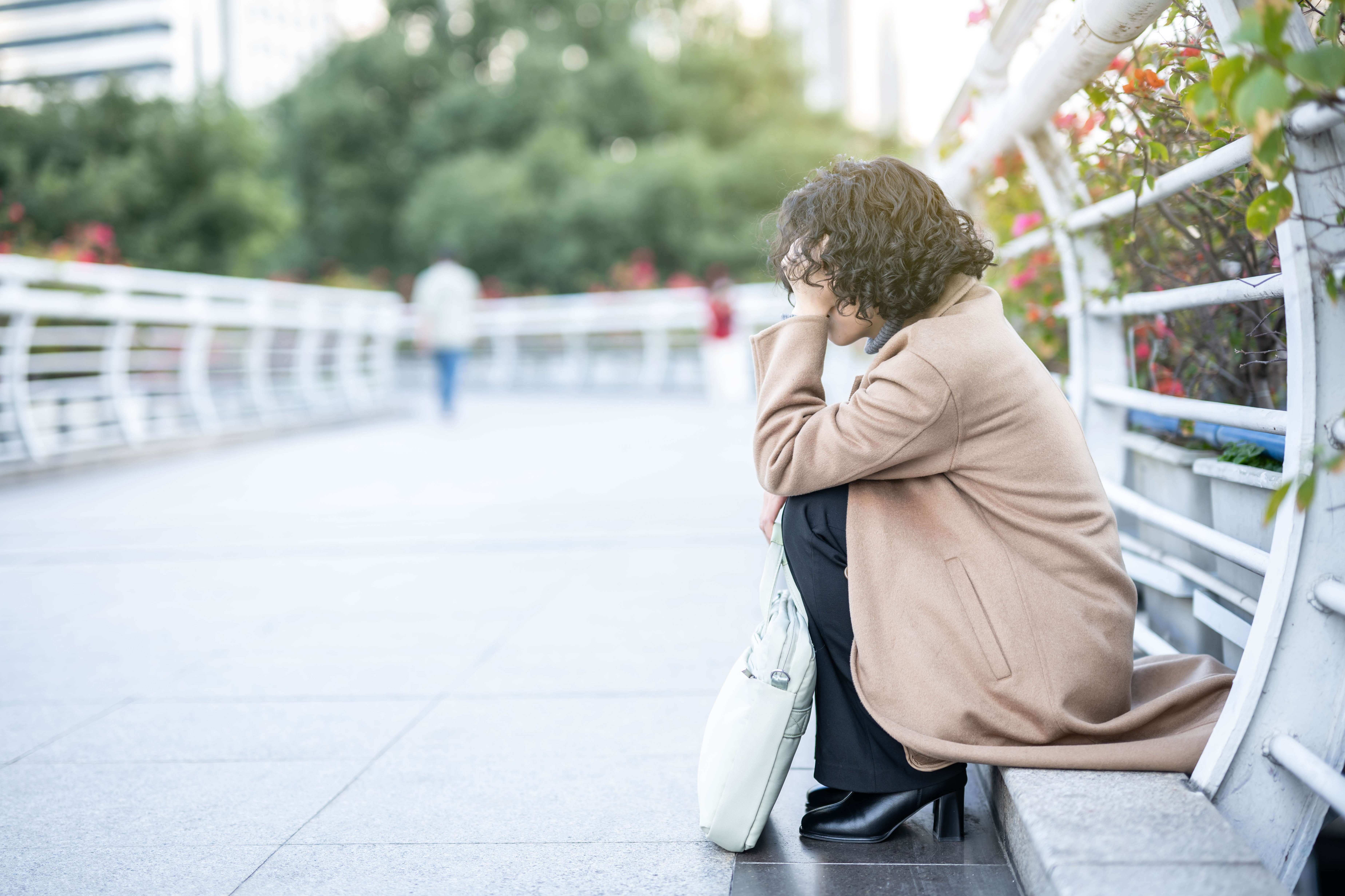A woman in a long beige coat is sitting on the ground of what looks like a bridge for pedestrians in a city; her head is in her hands.