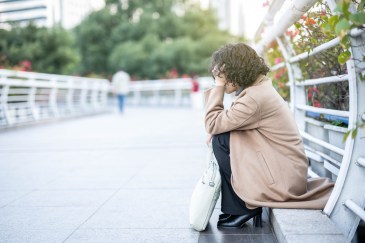 A woman in a long beige coat is sitting on the ground of what looks like a bridge for pedestrians in a city; her head is in her hands.