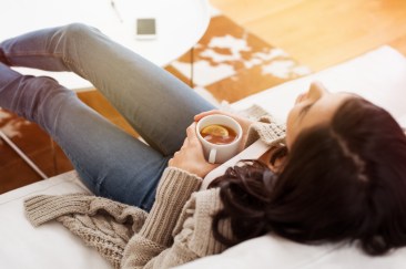 A woman in jeans and a light brown cardigan is lying back on her white couch, looking up with a mug of hot tea in her hands.