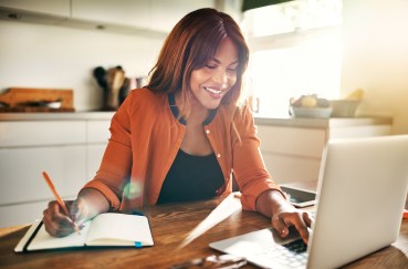 Woman sitting at kitchen table working on laptop, smiling