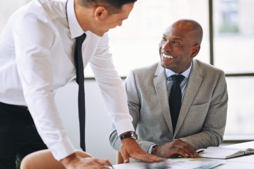 Two male employees talking at work, one seated, smiling