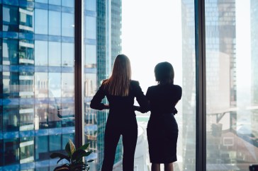 Two female colleagues stand side by side, looking out of a glass skyscraper into the rest of the city during the day.