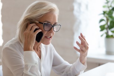 A white woman in her 50s is talking angrily on the iphone at her white desk during the day.