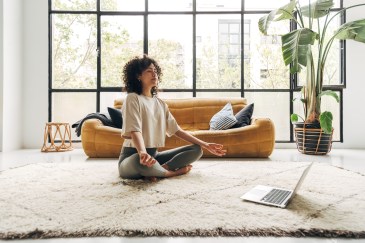 A Black woman is meditating in her living room with her laptop in front of her; there's a big window behind her with daylight streaming through it and an orange couch.