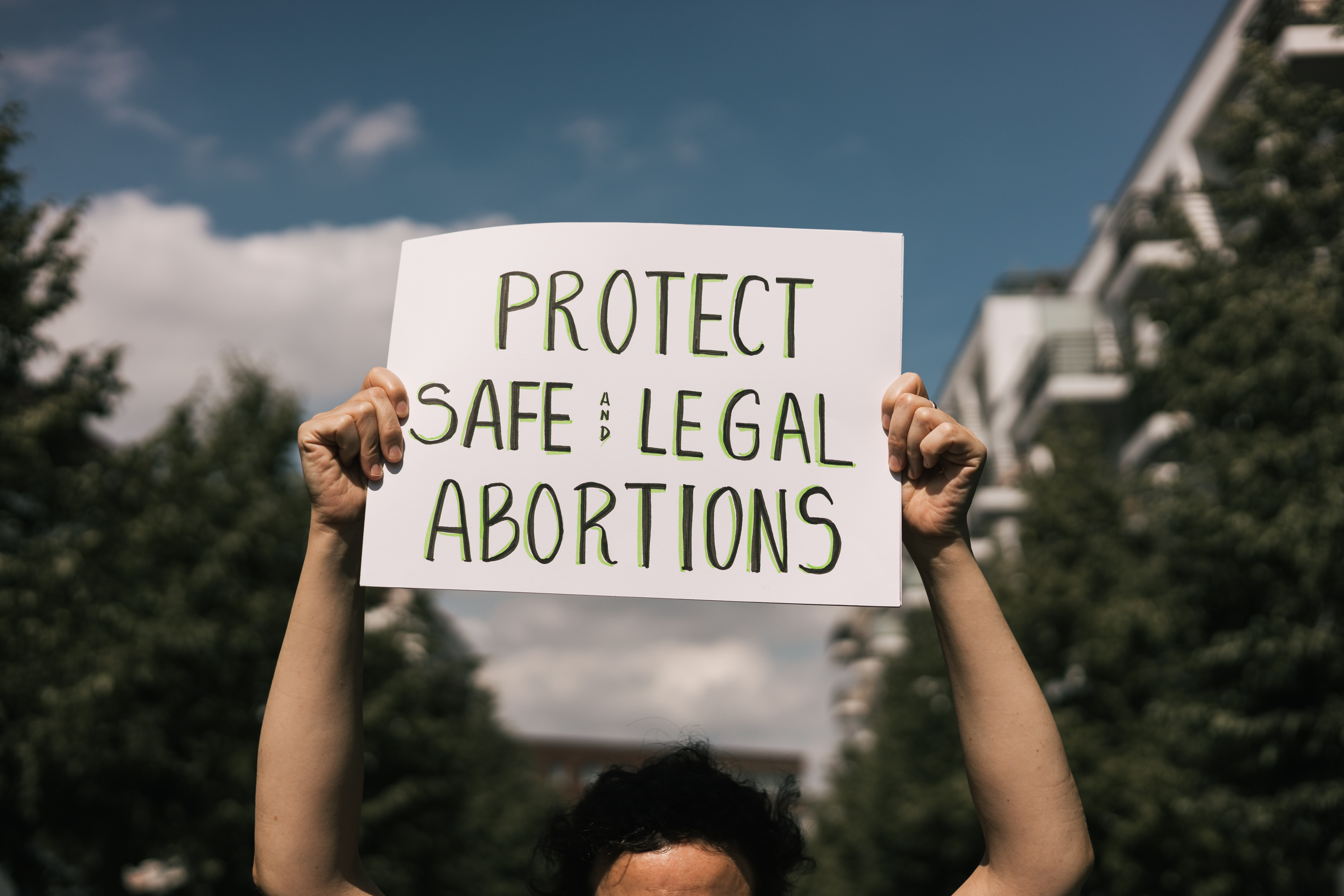A woman holding a sign that says, "Protect safe and legal abortions" outside on a street lined with green trees. Only the sign is in focus.