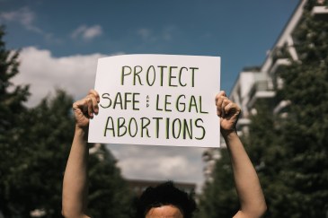 A woman holding a sign that says, "Protect safe and legal abortions" outside on a street lined with green trees. Only the sign is in focus.