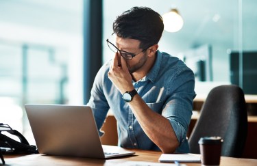 A man with glasses is pinching the bridge of his nose in frustration while sitting in front of his laptop.